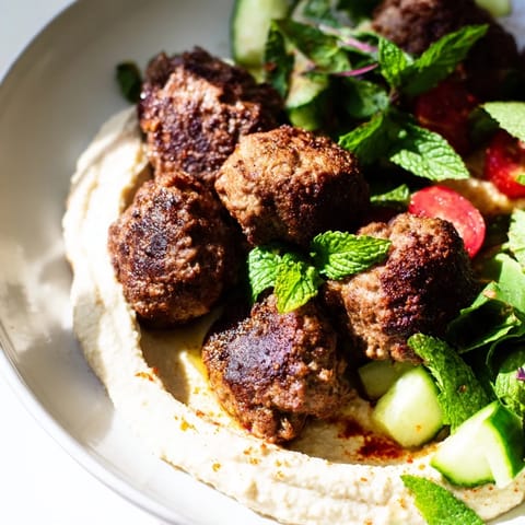 A close-up of venison meatballs, their spiced surface glistening, served over a bed of fresh salad and hummus.